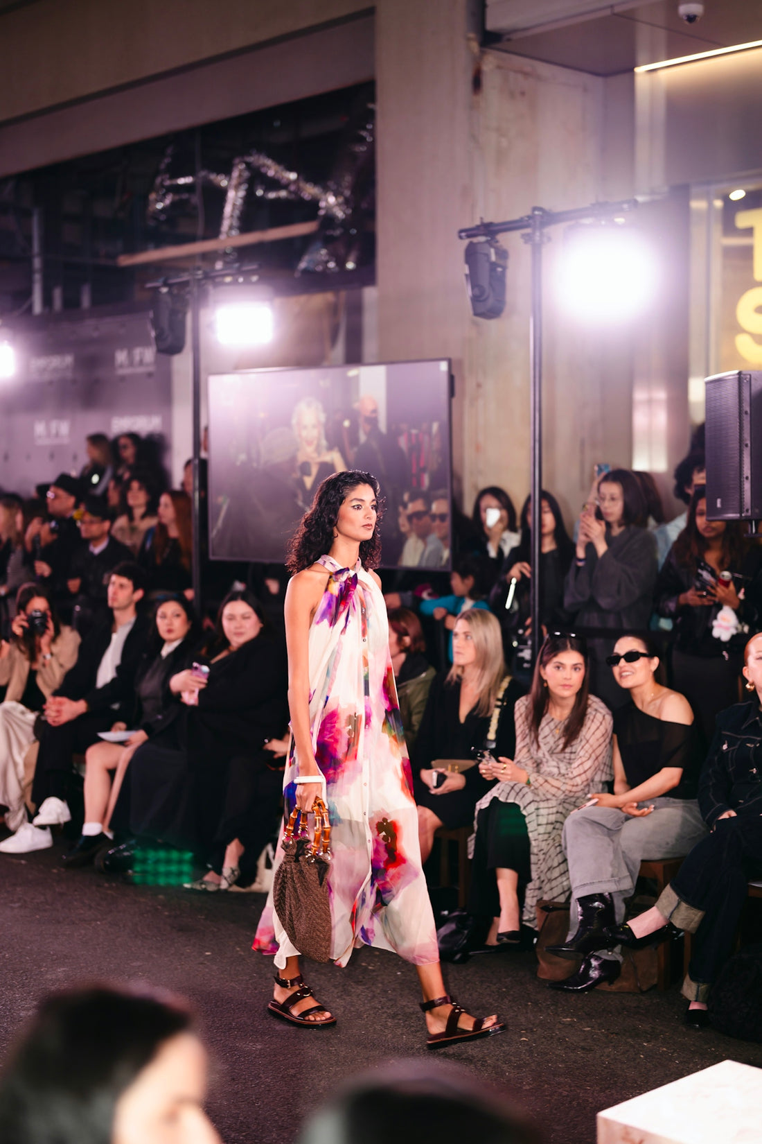 Model walks runway in floral dress with audience watching