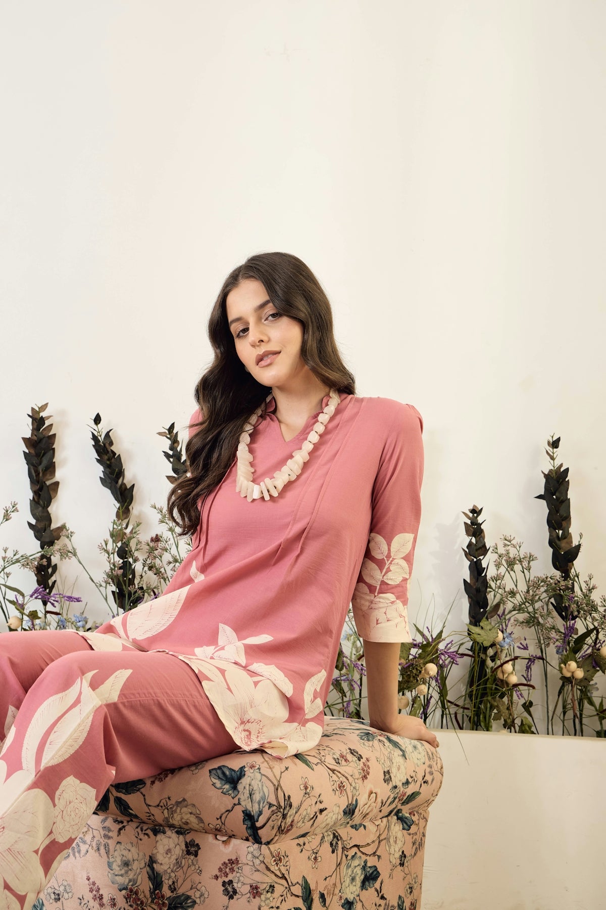 Woman in pink floral outfit sits near dried flowers