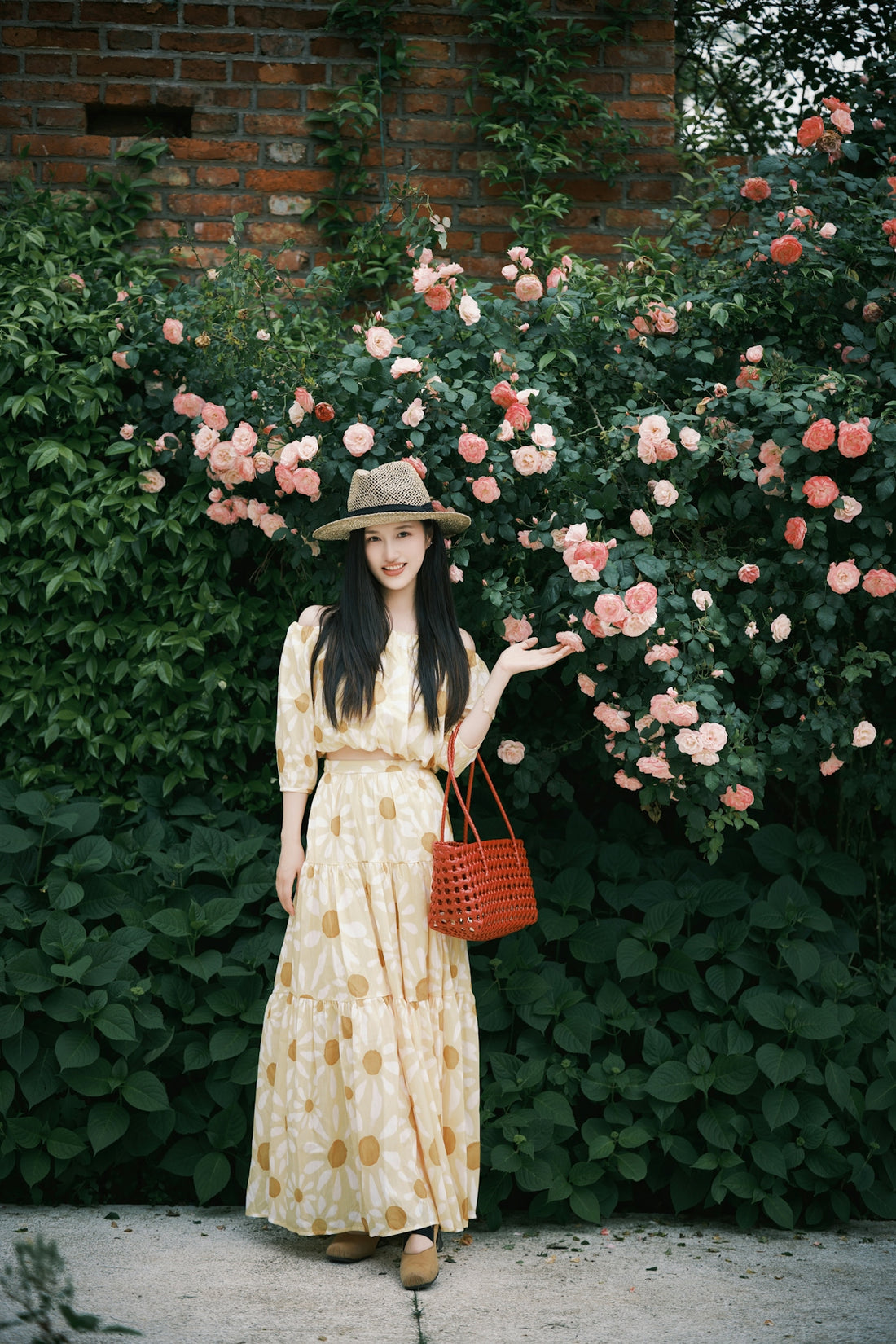 A woman in a hat and dress stands by roses