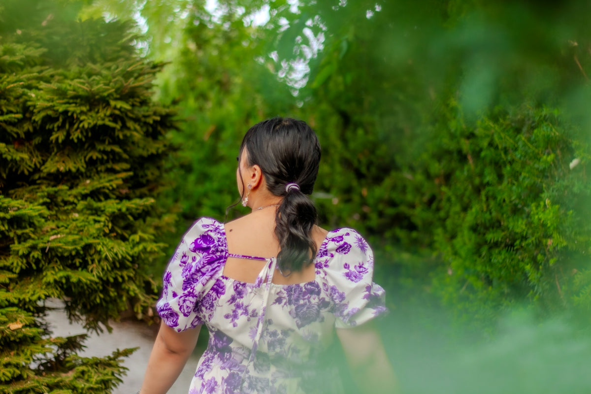A woman in a white dress with purple floral patterns walking through green trees.