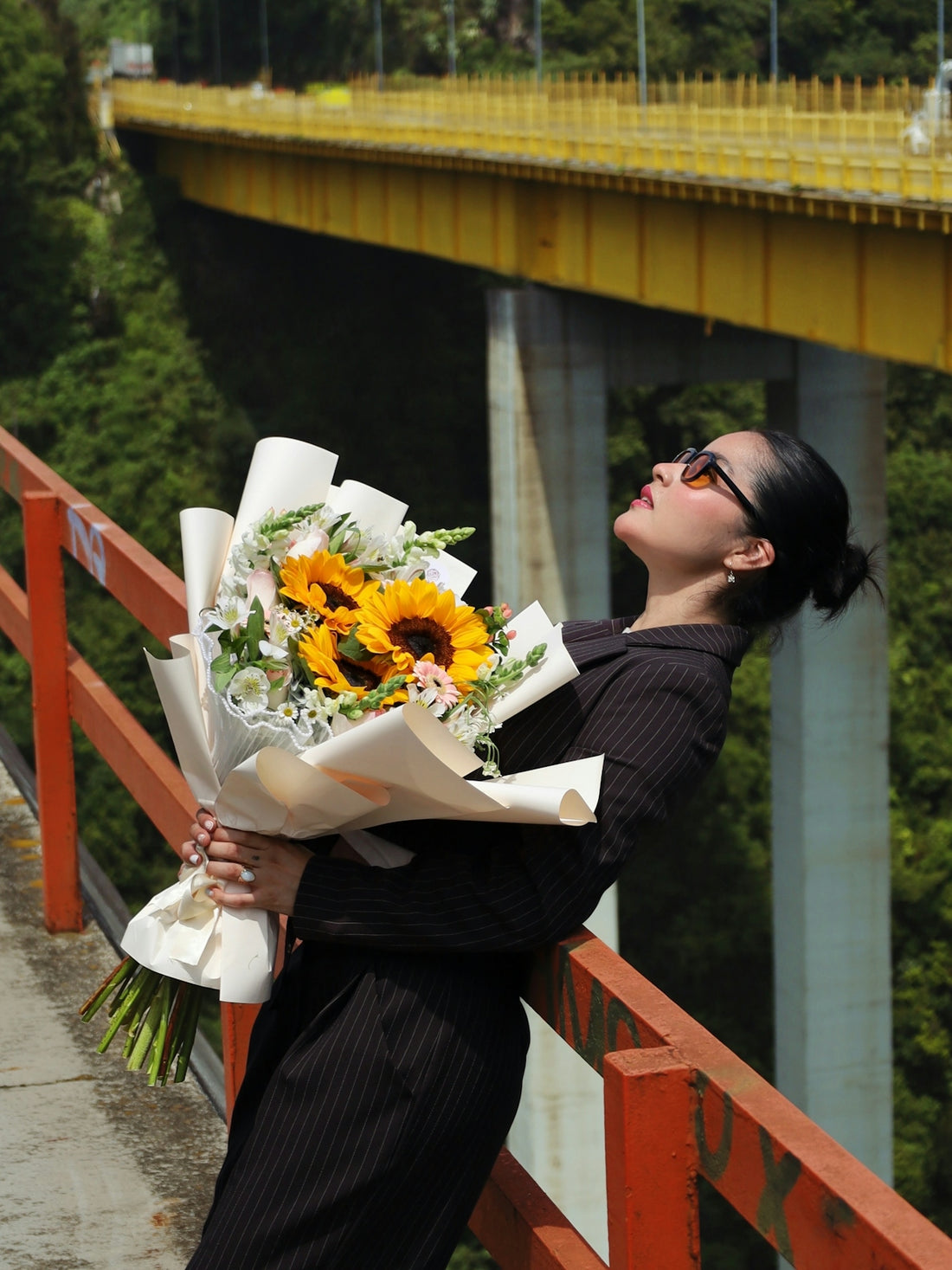 A woman in a pinstripe blazer holding a sunflower bouquet on a bridge